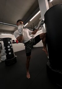 Boxo treniņš, kurā redzams cilvēks ar sudraba boksa cimdiem, baltu krekliņu un melniem šortiem, sitot melno boksa maisu.