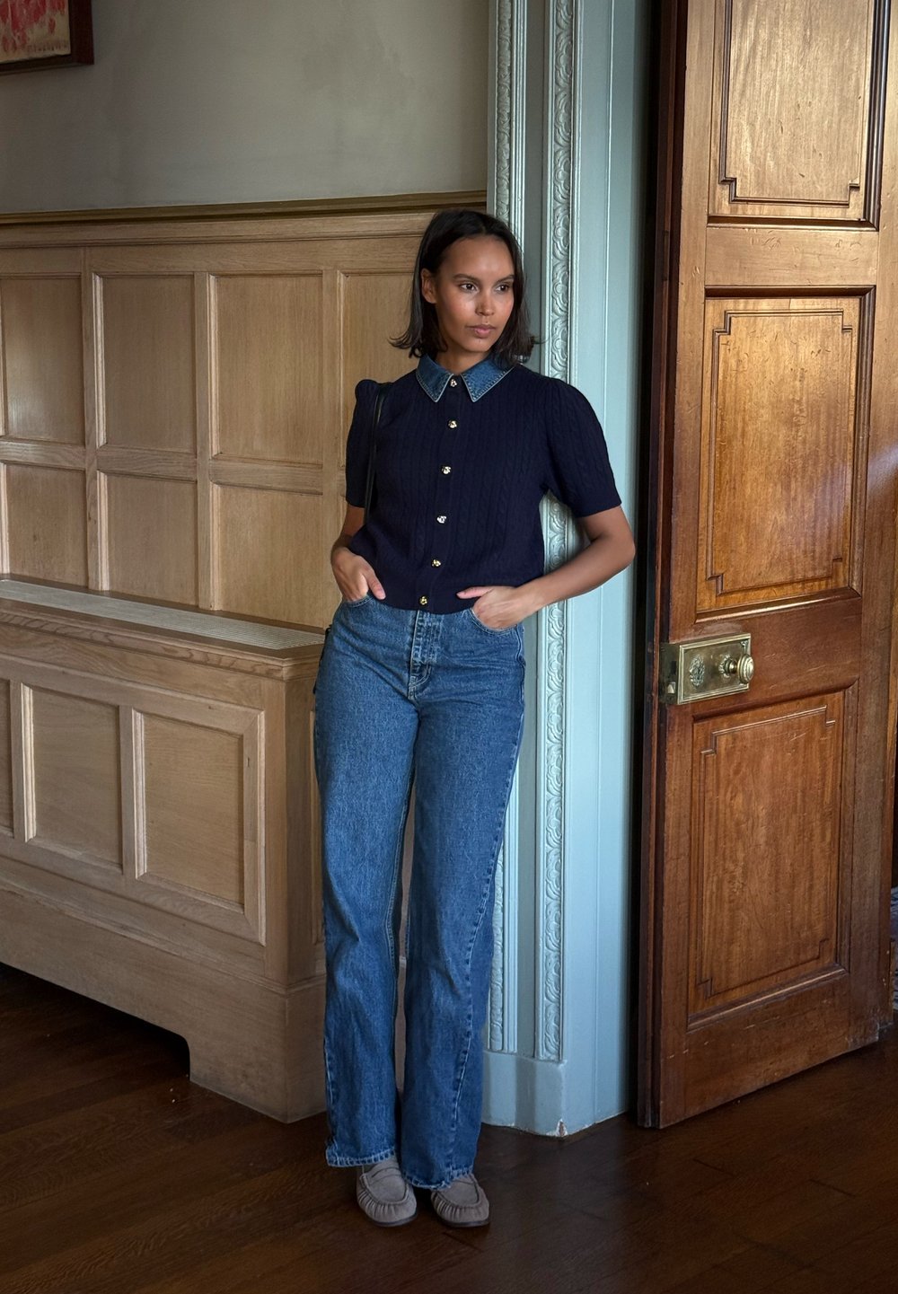 Chemise bleu marine à manches courtes en maille torsadée avec col en denim, boutons dorés, associée à un jean bleu taille haute et des ballerines grises, sur un fond en lambris de bois.