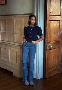 Chemise bleu marine à manches courtes en maille torsadée avec col en denim, boutons dorés, associée à un jean bleu taille haute et des ballerines grises, sur un fond en lambris de bois.