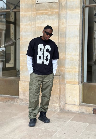 Hombre con gafas de sol negras, camiseta negra con estampado blanco "86" y estrella sobre mangas largas blancas, pantalones cargo oliva y botas negras, de pie al aire libre.