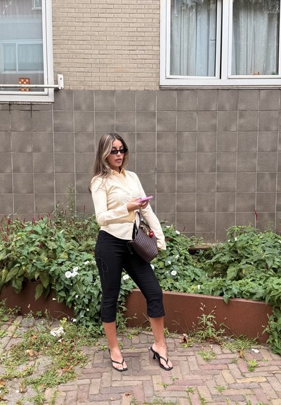 Camisa de botones a rayas crema, pantalones negros cortos con lazadas laterales, sandalias negras, sosteniendo un teléfono rosa, de pie junto a plantas verdes.