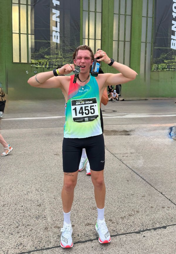 Coureur portant le dossard 1455 tient une médaille et une boisson, célébrant après avoir terminé une course de 10 km en plein air près de bâtiments verts.