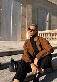 Hnědá semišová bunda s černým rolákem, černými kalhotami a slunečními brýlemi. Sedí na kamenných schodech a vedle ní je kabelka. Pozadí osvětlené sluncem.