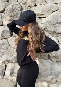 Gorra negra con un logo, combinada con una sudadera negra con capucha que presenta un estampado dorado. Cabello largo y ondulado parcialmente visible. Leggings negros.
