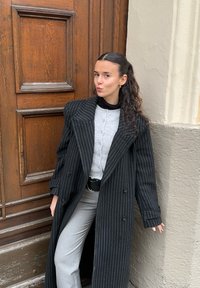 Lungo cappotto nero a righe sottili sopra una camicia grigia con bottoni e pantaloni grigi, completato da una cintura nera. Texture lisce con modelli distintivi.
