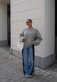 Suéter de punto gris claro de gran tamaño con mangas anchas, vaqueros azules con rayas laterales verdes, zapatos beige y un bolso de mano tejido blanco.