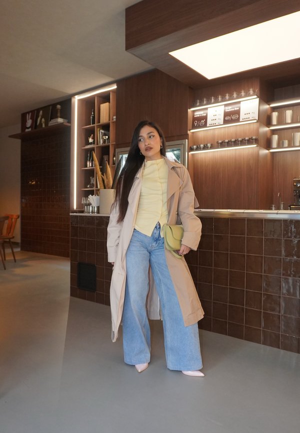 Kobieta z długimi ciemnymi włosami, ubrana w beżowy trencz, żółty top, szerokie dżinsy i trzymająca zieloną torbę we wnętrzu nowoczesnej kawiarni.