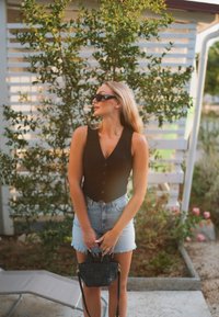 Black sleeveless button-up top, high-waisted denim shorts, holding a small black woven handbag. The background showcases lush green foliage.