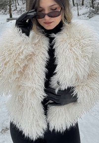 Femme portant un manteau blanc duveteux, des gants noirs et des lunettes de soleil étroites dans un décor extérieur enneigé.