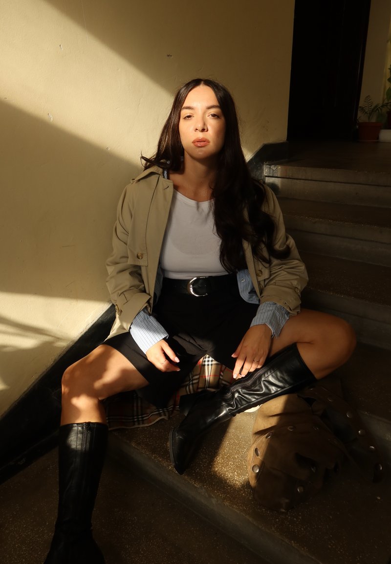 Trench coat beige chiaro sopra una camicia bianca, gonna nera e stivali neri fino al ginocchio. Seduta sui gradini con una borsa marrone e tessuto scozzese.