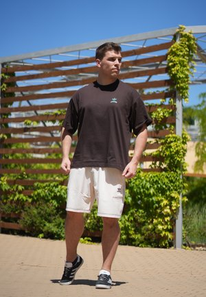 Joven con una camiseta negra, pantalones cortos blancos y zapatillas negras caminando al aire libre cerca de una pérgola de madera cubierta de enredaderas verdes.