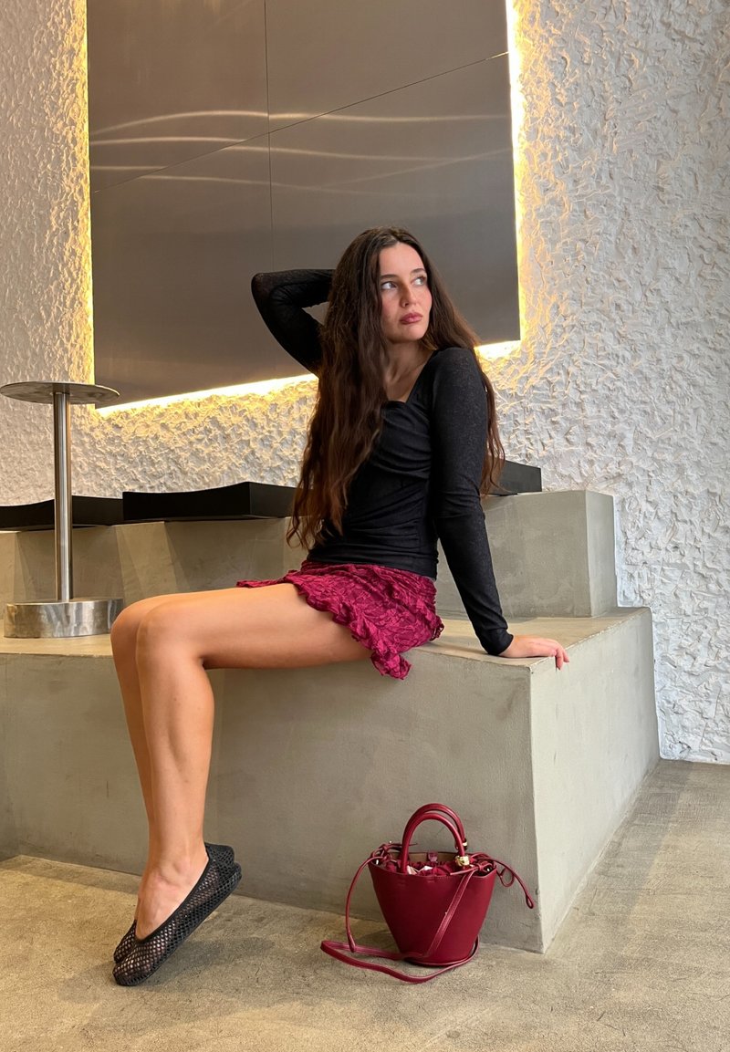 Schwarzes Langarmoberteil, rosa Rüschensrock, schwarze Netzflats und eine bordeauxfarbene Bucket Bag auf einer strukturierten Betonplattform mit sanfter Beleuchtung.