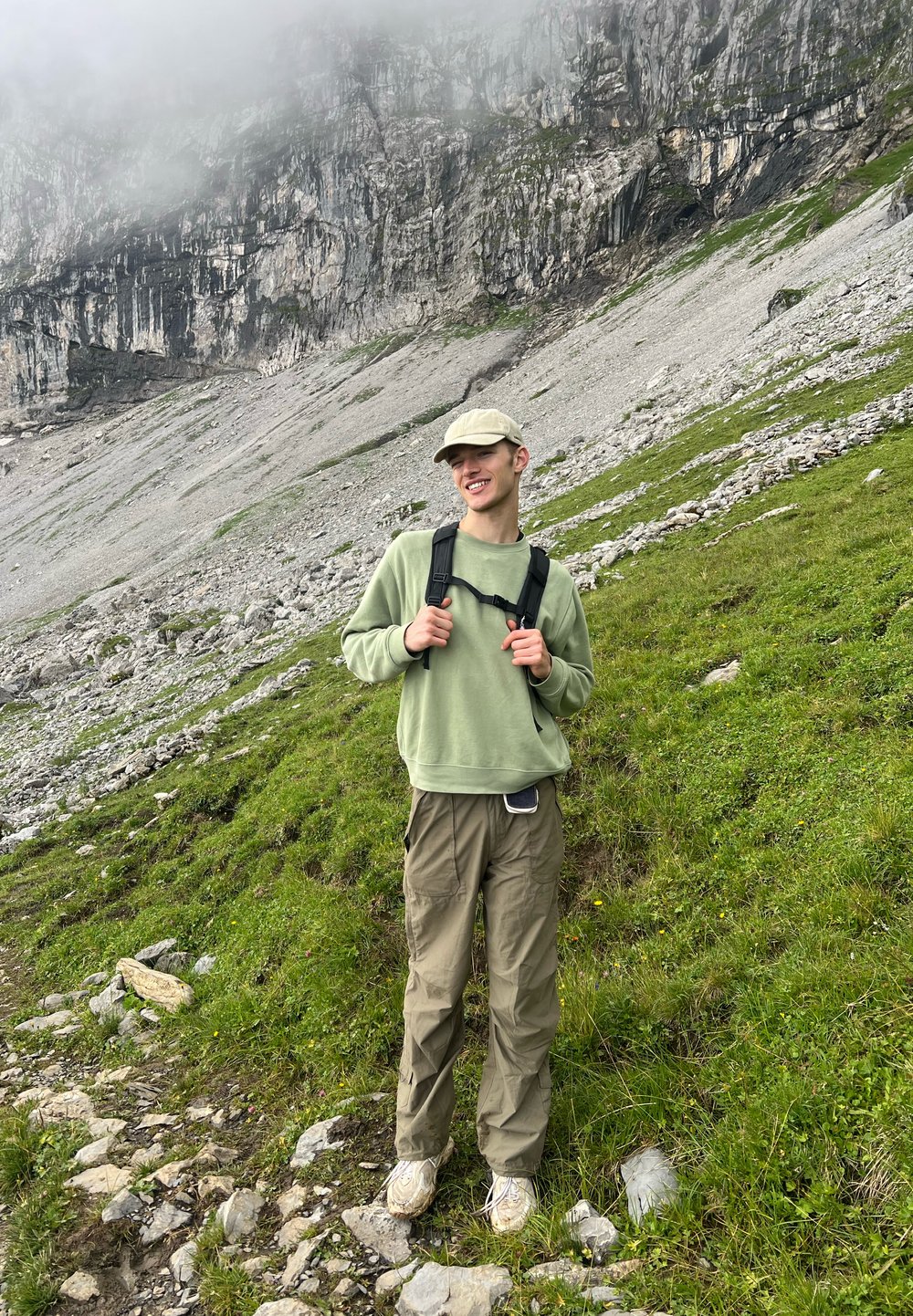 Mladý člověk v čepici, zelené mikině a turistických kalhotách stojí na travnatém, skalnatém svahu hory s mlhavými útesy v pozadí.