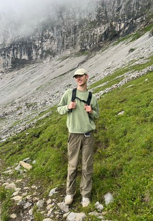 Joven con gorra, sudadera verde y pantalones de senderismo se encuentra en una ladera montañosa de hierba y rocas, con acantilados neblinosos de fondo.