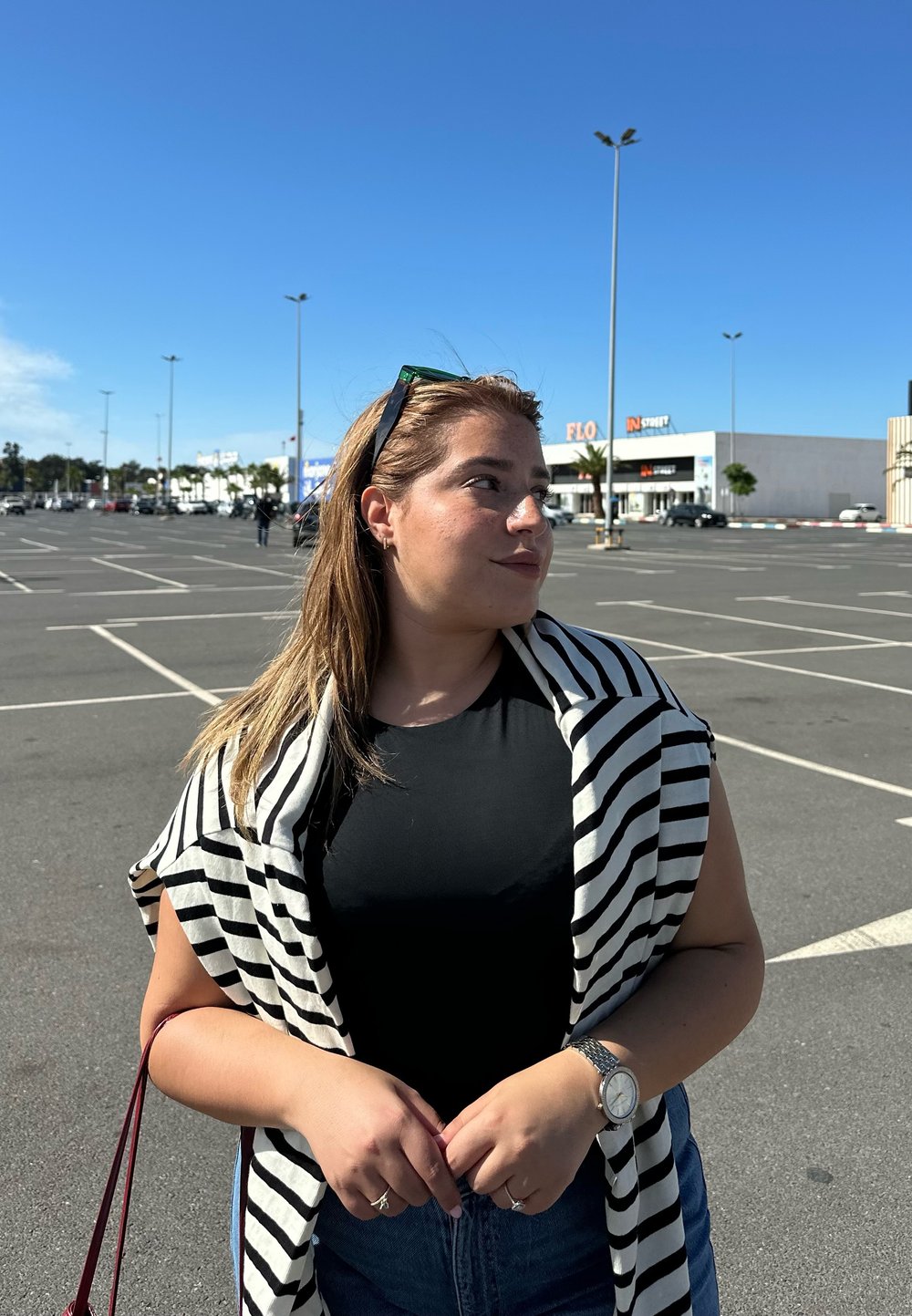 T-shirt nera, sciarpa a strisce bianche e nere, e jeans di denim. Orologio argento al polso. Ambientazione all'aperto con cielo blu e sfondo di un parcheggio.