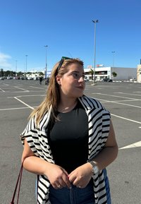 T-shirt nera, sciarpa a strisce bianche e nere, e jeans di denim. Orologio argento al polso. Ambientazione all'aperto con cielo blu e sfondo di un parcheggio.