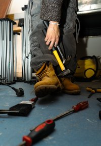 Žlté pracovné topánky, šedé nohavice a textúrovaný hnedý sveter; drží kladivo a rôzne nástroje sú rozptýlené po modrej podlahe.