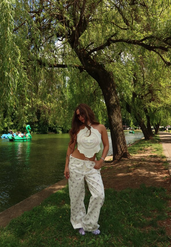 Donna con occhiali da sole che indossa un top bianco all'uncinetto e pantaloni floreali in piedi accanto a un fiume fiancheggiato da salici e pedalò in una giornata di sole.
