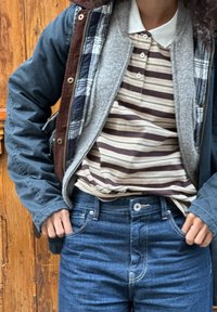 Personne portant un jean bleu, un polo rayé, une polaire grise superposée et une veste à carreaux, debout avec les mains dans les poches contre une porte en bois.