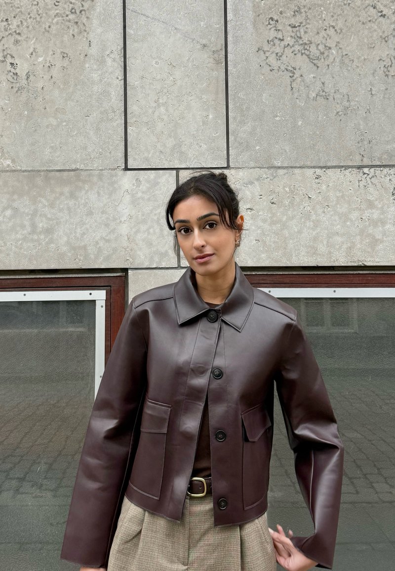 Giacca corta in pelle marrone con collo a risvolto, chiusura con bottoni e due tasche frontali, abbinata a pantaloni beige a quadri.