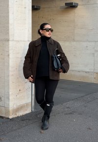 Brun semsket jakke over en svart høyhalset genser, kombinert med svarte leggings og svarte gummistøvler. Holder en liten svart håndveske.