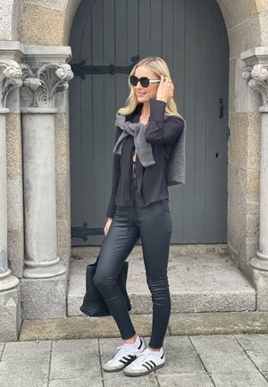 Top negro de manga larga con detalle de lazo, suéter gris drapeado por encima, pantalones de imitación cuero negros, bolsa trenzada negra, zapatillas blancas con rayas negras.