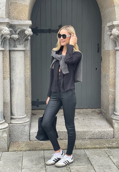 Top negro de manga larga con detalle de lazo, suéter gris drapeado por encima, pantalones de imitación cuero negros, bolsa trenzada negra, zapatillas blancas con rayas negras.