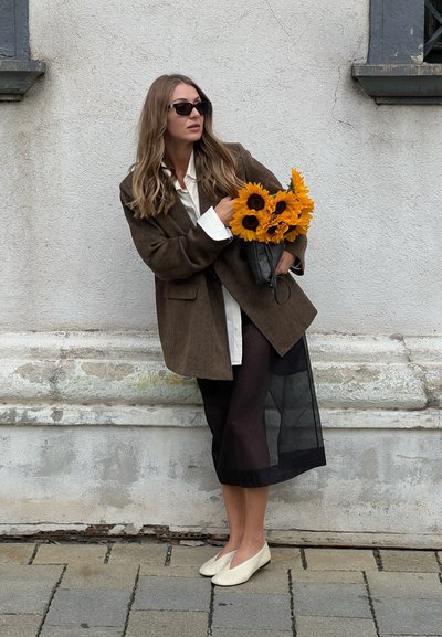 Blazer marrón oversized sobre una camisa blanca, falda negra transparente y bailarinas crema. Sostenido un ramo de girasoles. Fondo urbano.
