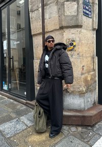 Črn puffer jakna, grafična črna-bela majica, črni hlače s pinstripi. Temna sončna očala, srebrni pas, zelena torba, urbana kamnita kulisa.