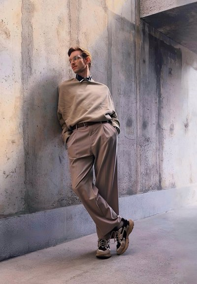 Suéter beige de gran tamaño superpuesto a una camisa negra con cuello, pantalones de tono marrón claro con pliegues y zapatillas negras y beige; de pie contra una pared de concreto.