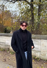 Μαύρο παλτό και μαύρο φούτερ με φερμουάρ πάνω α�πό μπλε τζιν. Τα αξεσουάρ περιλαμβάνουν κόκκινα γυαλιά ηλίου. Το φόντο περιλαμβάνει πέτρινο τοίχο και φθινοπωρινά φύλλα.