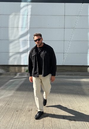 Camisa negra abotonada sobre una camiseta negra, pantalones beige y zapatos negros, caminando sobre una superficie gris texturizada con una pared perforada al fondo.