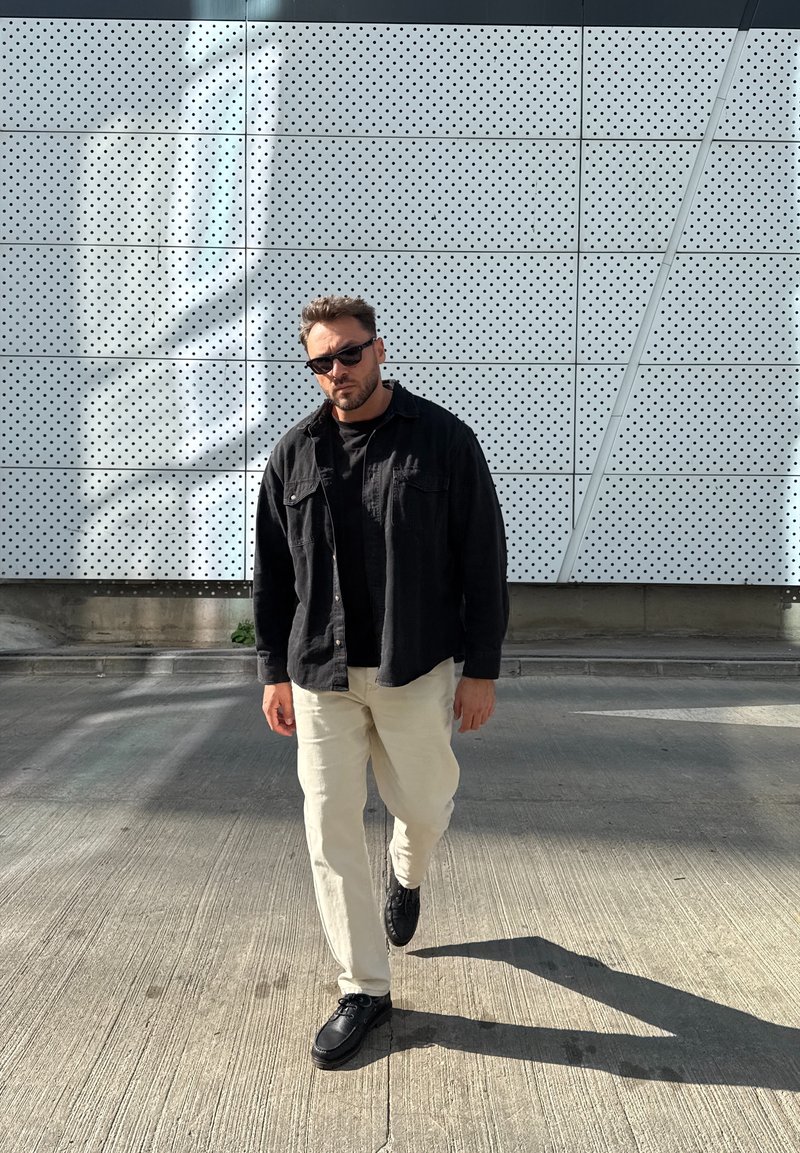 Čierna košeľa na gombíky cez čierne tričko, béžové nohavice a čierne topánky, kráčajúci po textúrovanom sivom povrchu s perforovanou stenou vzadu.