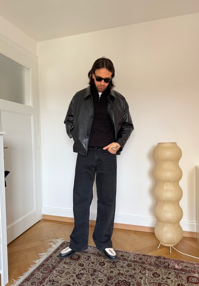 Hombre con chaqueta de cuero negra, pantalones negros y gafas de sol, de pie sobre una alfombra con estampado en una habitación minimalista con una lámpara de pie escultórica beige.