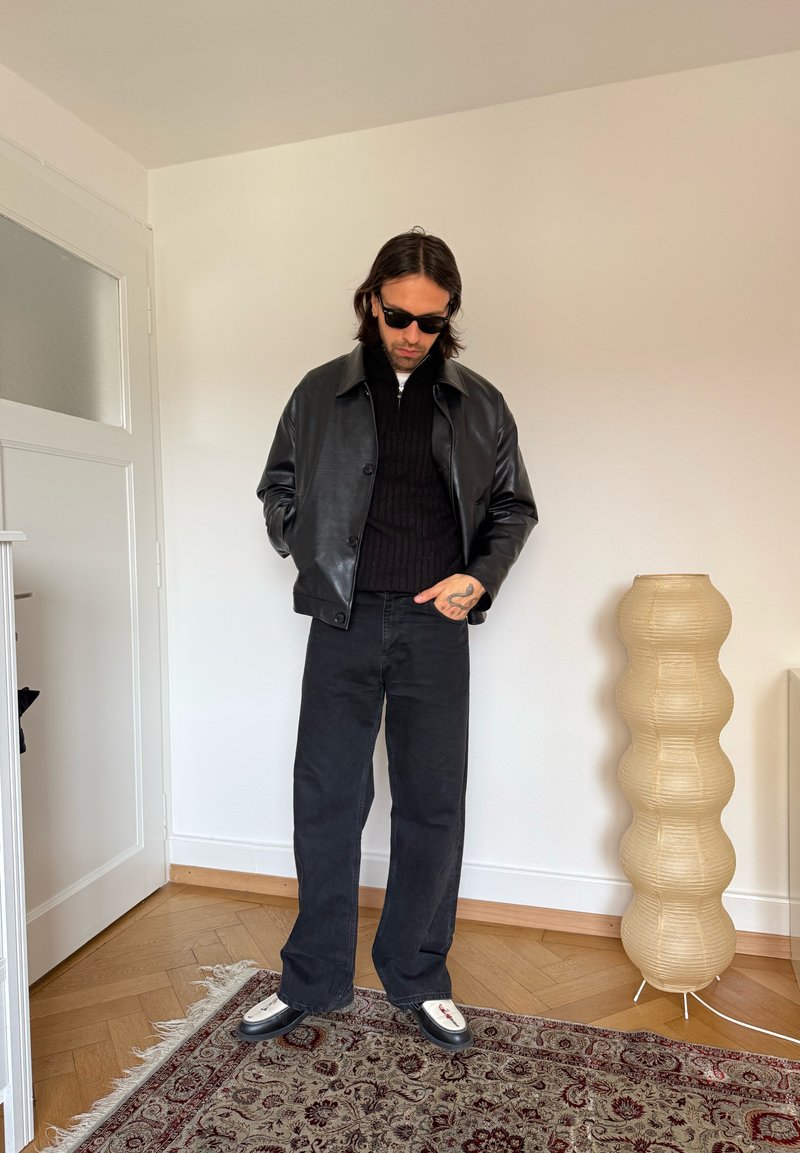 Man draagt een zwarte leren jas, zwarte broek en zonnebril en staat op een patroonrijke tapijt in een minimalistische kamer met een sculpturale beige vloerlamp.