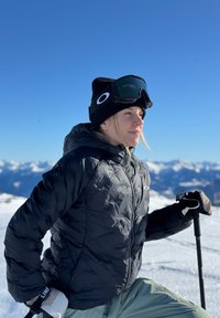 Casaco puffer preto com padrão de estrelas, calças de esqui verdes, gorro preto e óculos de esqui. Fundo de montanha nevada sob um céu azul claro.