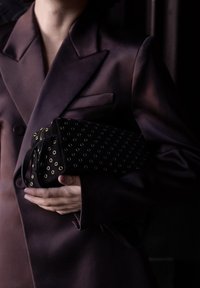 Personne portant un blazer violet foncé tenant un sac noir avec des  œillets dorés sur un fond sombre.