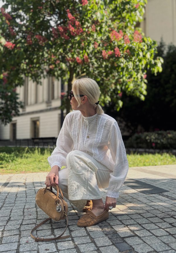Femme aux cheveux blonds attachés porte un chemisier en dentelle blanc et un pantalon, des mocassins marron et des lunettes de soleil, accroupie sur un chemin pavé près d’un arbre.