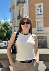 Valkoinen hihaton toppi mustilla muotoiluilla, beige korkeavyötäröiset housut, musta käsilaukku ja kilpikonnakuosiset aurinkolasit. Taustalla kirkas sininen taivas.