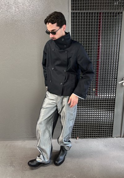 Hombre con chaqueta negra abotonada, pantalones metálicos plateados, botas negras y gafas de sol, de pie frente a una pared gris y una puerta de rejilla metálica.