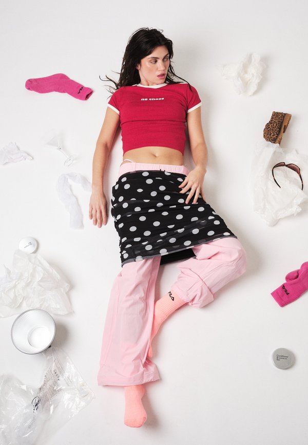 Ung kvinne i rosa bukser og rød crop-top ligger på gulvet omgitt av spredte sokker, et leopardmønstret sko, solbriller, papir og en bolle.