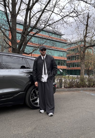 Chaqueta bomber negra sobre una camisa blanca, corbata negra y pantalones grises holgados. De pie junto a un SUV oscuro, con edificios modernos de cristal de fondo.
