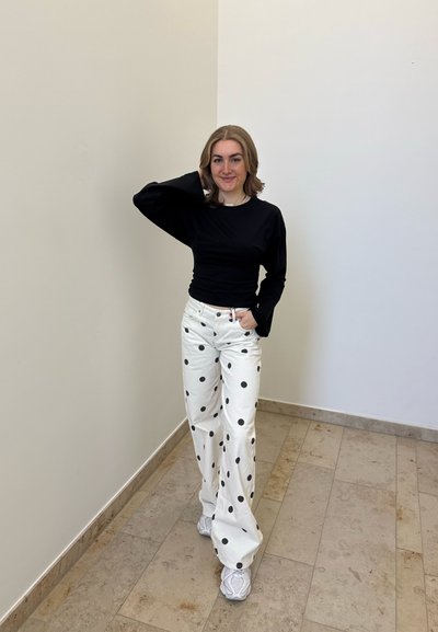 Mujer de pie en el interior, lleva una camiseta negra de manga larga, pantalones blancos con lunares negros y zapatillas blancas, sonriendo con una mano en la cabeza.