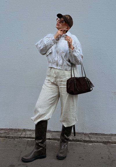 Chaqueta bomber de lino gris claro sobre pantalones de lino crema, botas altas marrón hasta la rodilla, bolso oscuro marrón y una gorra de béisbol marrón con logo.