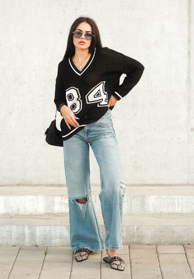 Suéter oversized de malla negra con el número blanco 84, combinado con unos jeans acampanados de color azul claro con rodillera desgastada y unos zapatos slip-on negros con estampado.