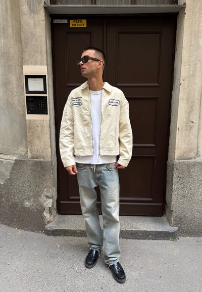 Chaqueta de mezclilla color crema con parches, camiseta blanca, jeans de un azul claro y zapatos negros. De pie frente a una puerta con una pared texturizada.