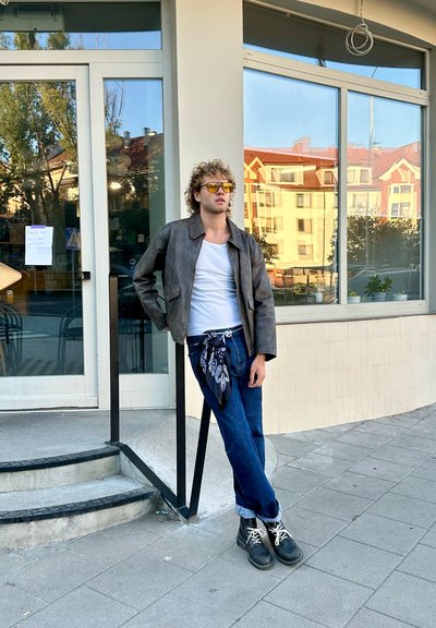 Chaqueta de cuero marrón, camiseta blanca, vaqueros azules con un pañuelo atado a la cintura y botas negras, de pie frente a un escaparate de cristal.