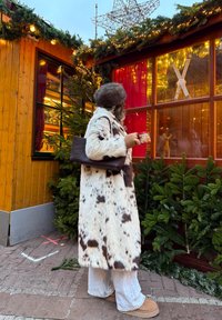 Manteau en fausse fourrure à motif tacheté, crème et marron, assorti à un pantalon blanc et des bottes marron ; tenant une tasse, dans une ambiance de marché festif.