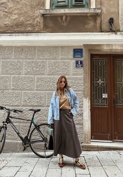 Chaqueta de mezclilla sobre una camisa marrón, combinada con una falda larga de cuero marrón. Bicicleta negra al lado, con un bolso azul texturizado y zapatos marrones.