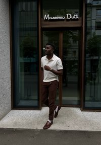 Krémové polo tričko, hnědé kordové kalhoty, hnědé boty s metalickým akcentem. Stojí u skleněných dveří, moderní městské prostředí.
